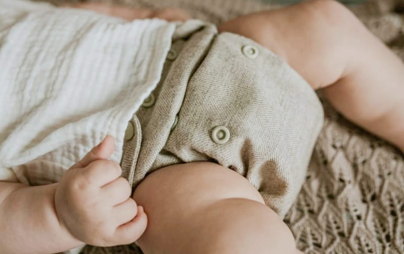 Een baby heeft een groen overbroekje van wol aan. Deze bevat drukknopen zodat het broekje makkelijk dicht gaat. De baby licht op zijn rug op een kleedje.